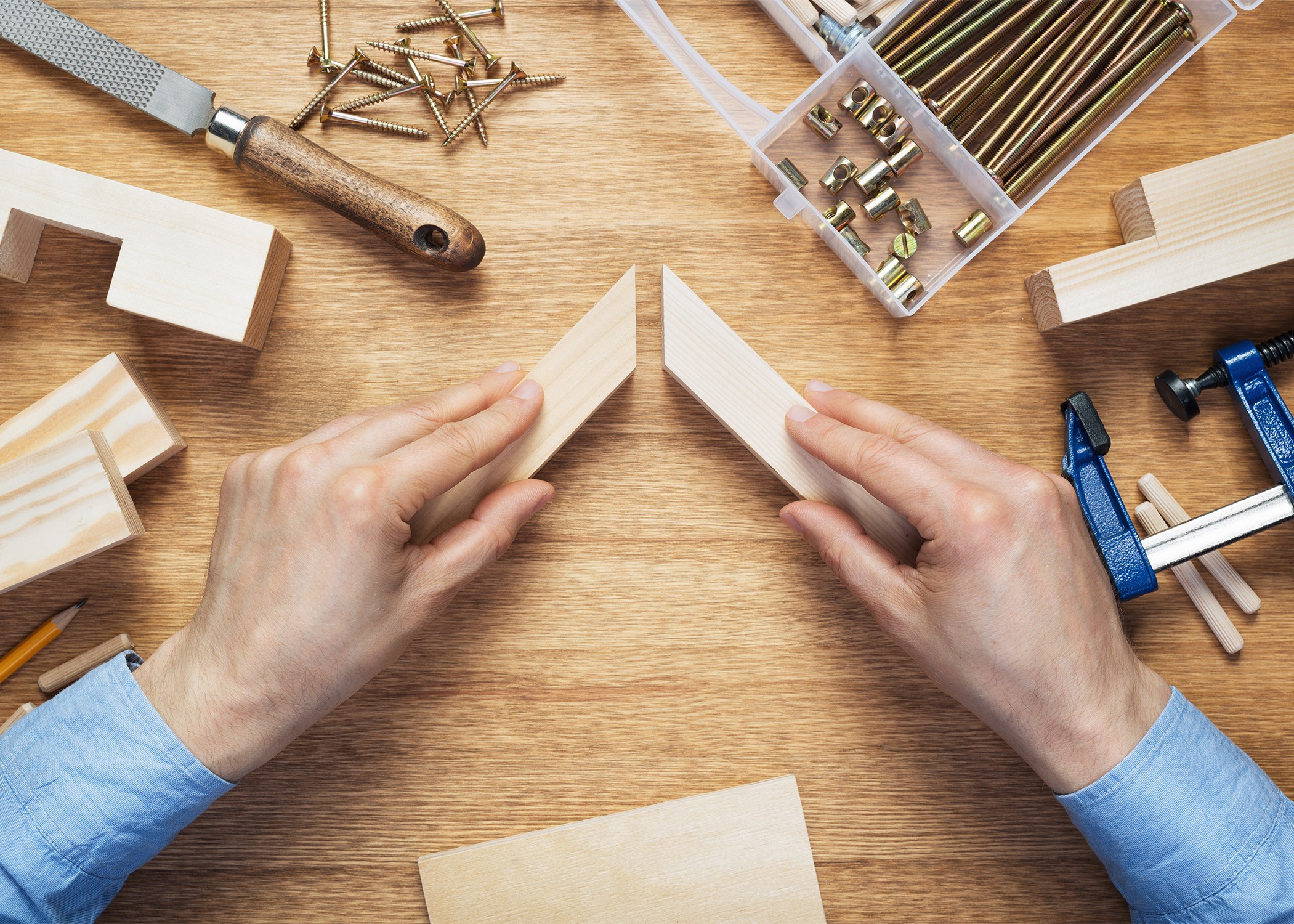 Hands in blue sleeves align two wooden pieces on a workbench surrounded by woodworking tools, screws, and clamps, conveying focus and craftsmanship.