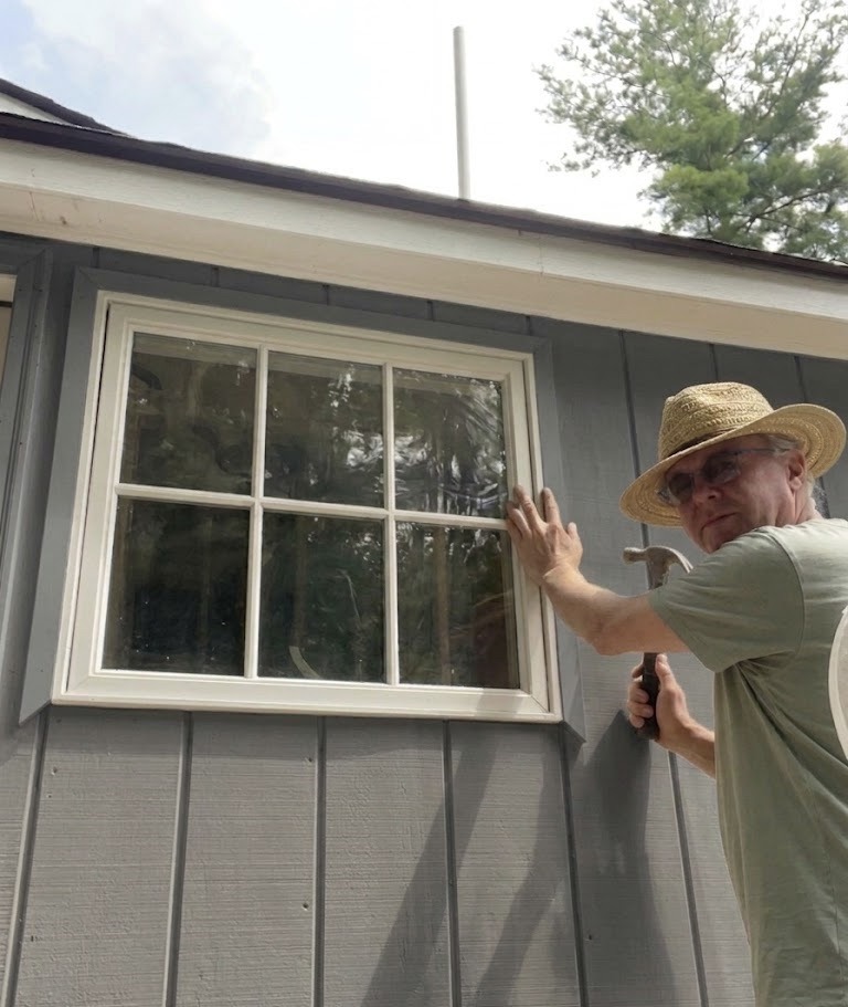 A person wearing a straw hat and glasses is repairing a window on a gray wooden building. They hold a hammer, conveying a focused, constructive mood.