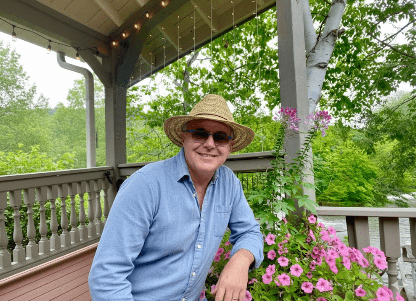 A man in a straw hat and sunglasses smiles while leaning on a railing adorned with pink flowers. He's on a porch overlooking lush green trees, exuding a relaxed, cheerful vibe.